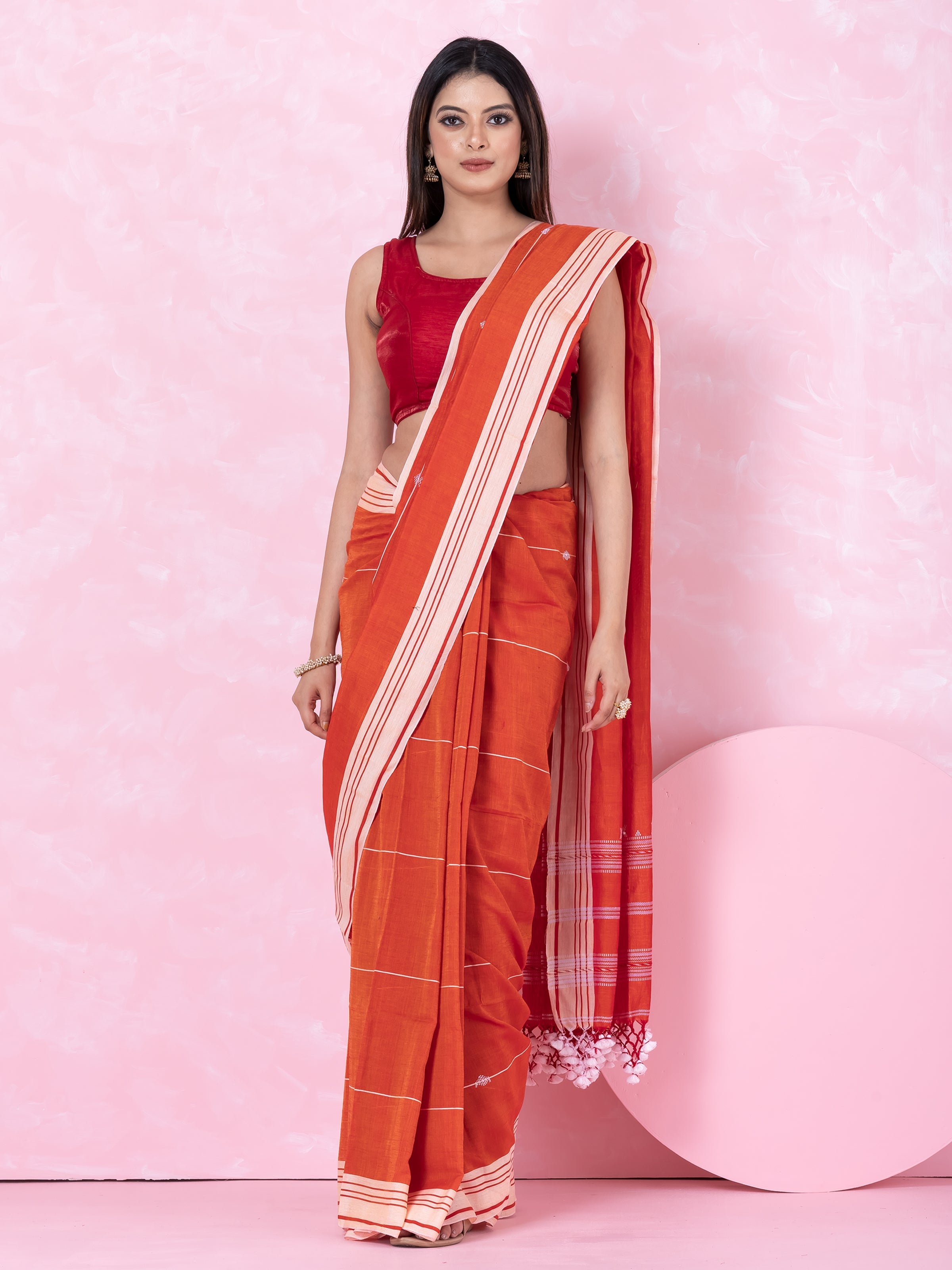 A woman modeling an orange- white handloom cotton saree with a white border, displayed against a pink background.