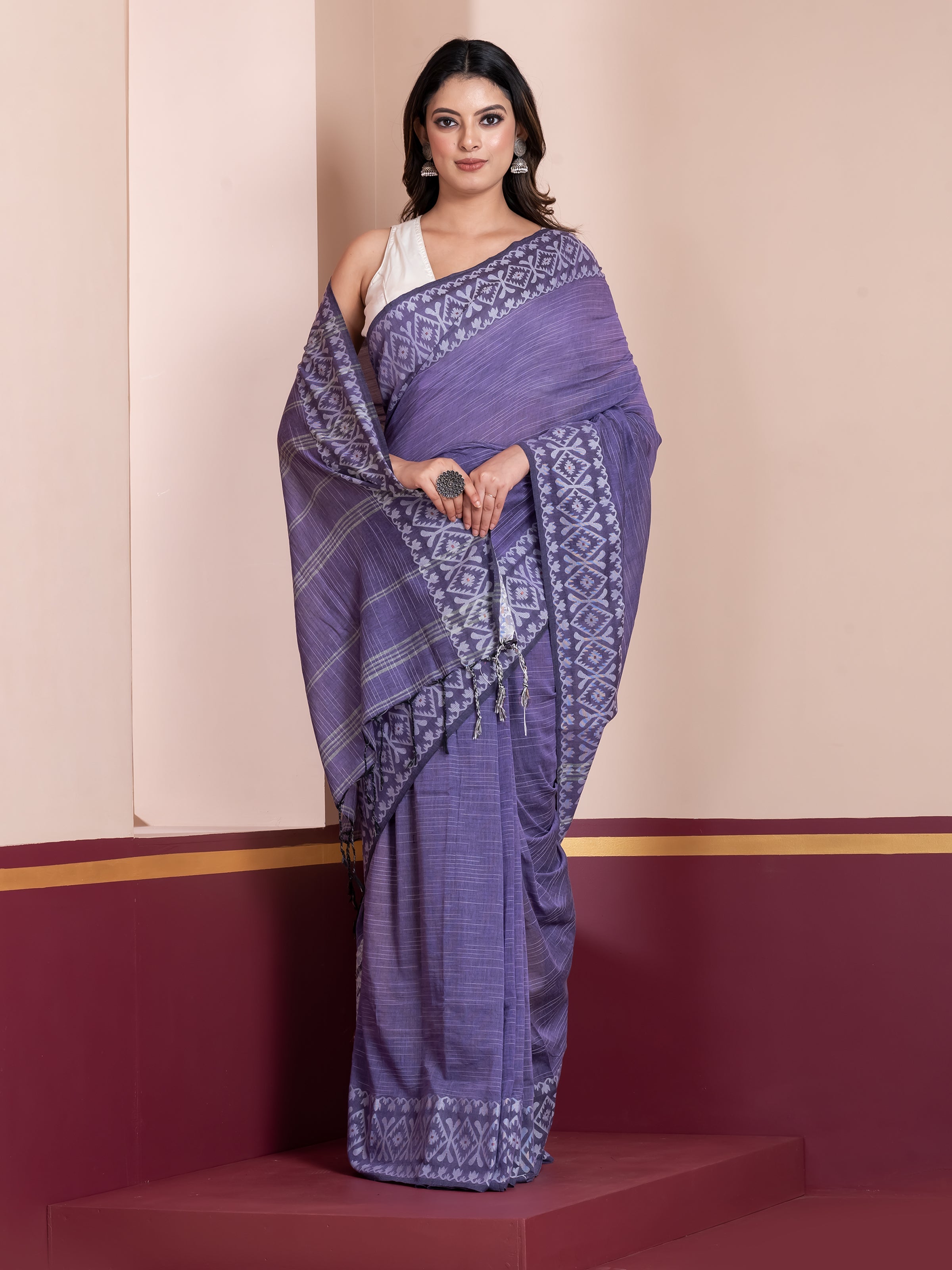 A woman modeling a violet khadi cotton saree with white border and geometric patterns, standing against a neutral background.