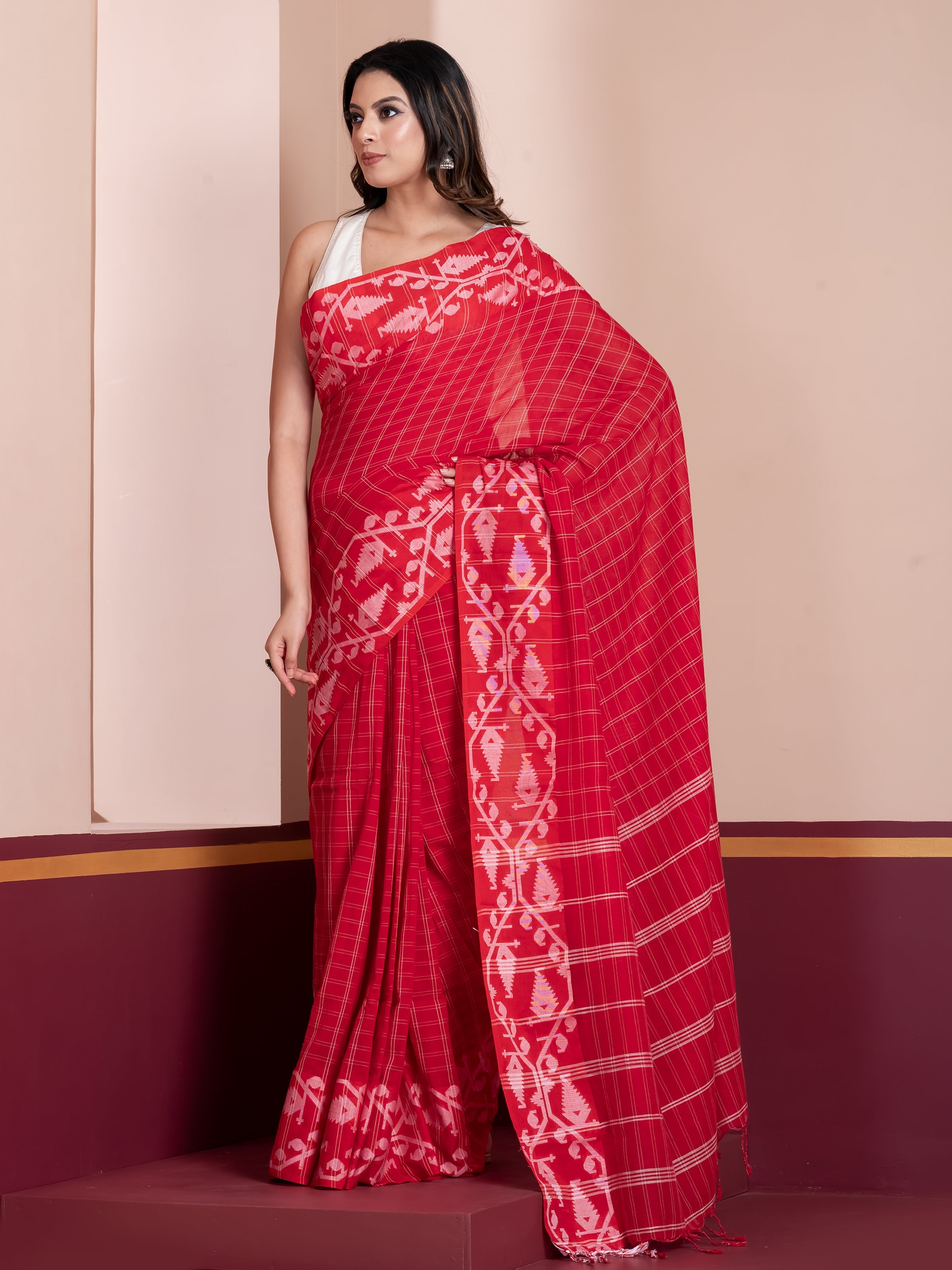 A woman modeling a red khadi cotton saree with white checks and Jamdani border patterns, standing against a beige background.