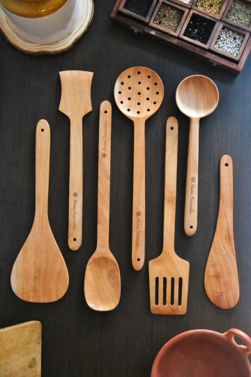 Seven piece neem wood kitchen utensil set by Almitra Sustainables arranged on dark surface including ladles, spatulas and stirring spoons, plastic-free handcrafted wooden cooking tools made from natural neem wood for sustainable kitchens.