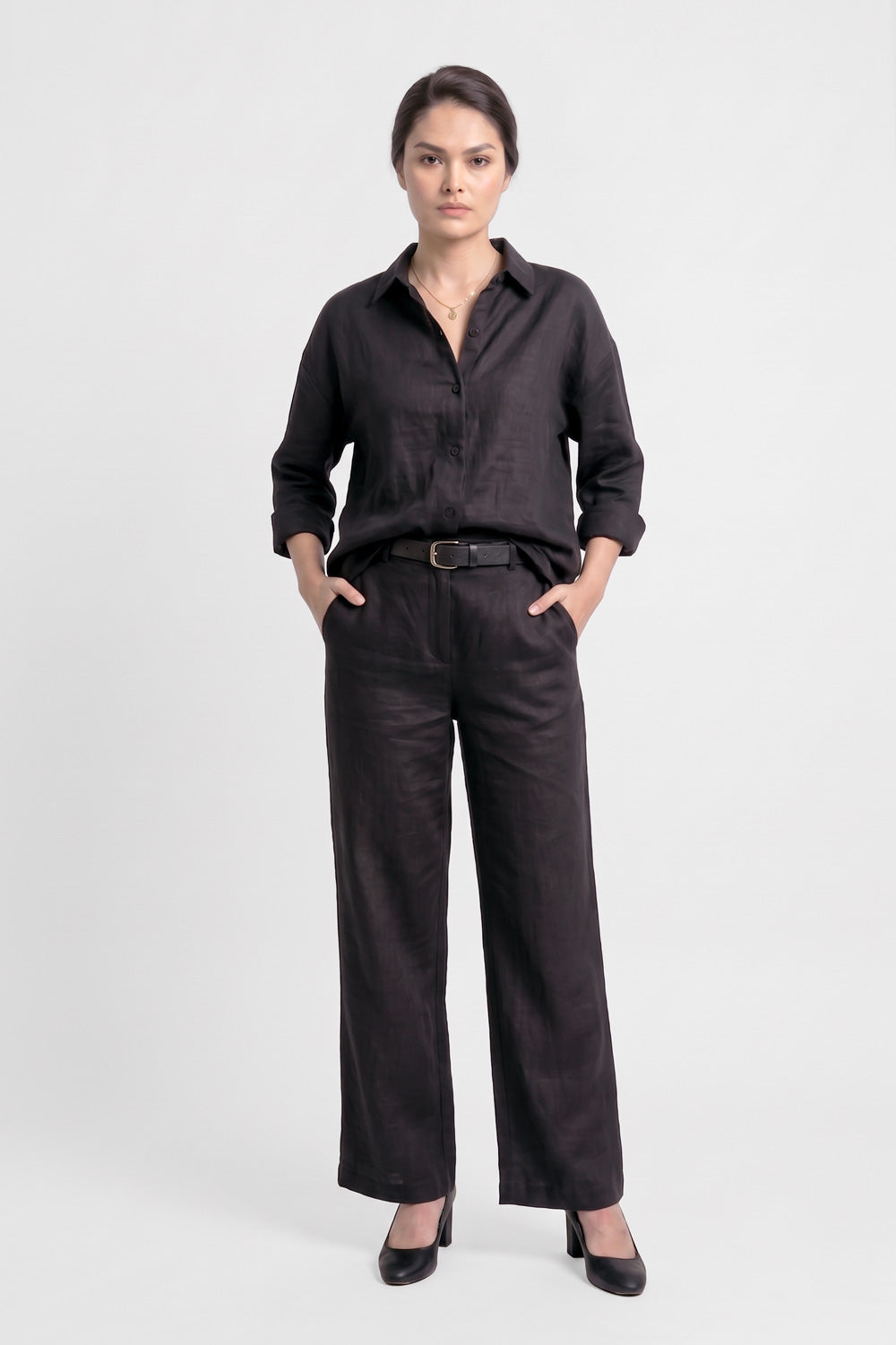 A model stands against a light gray studio background wearing a black long-sleeve button-up linen shirt, black wide-leg trousers, a black belt and black heels.