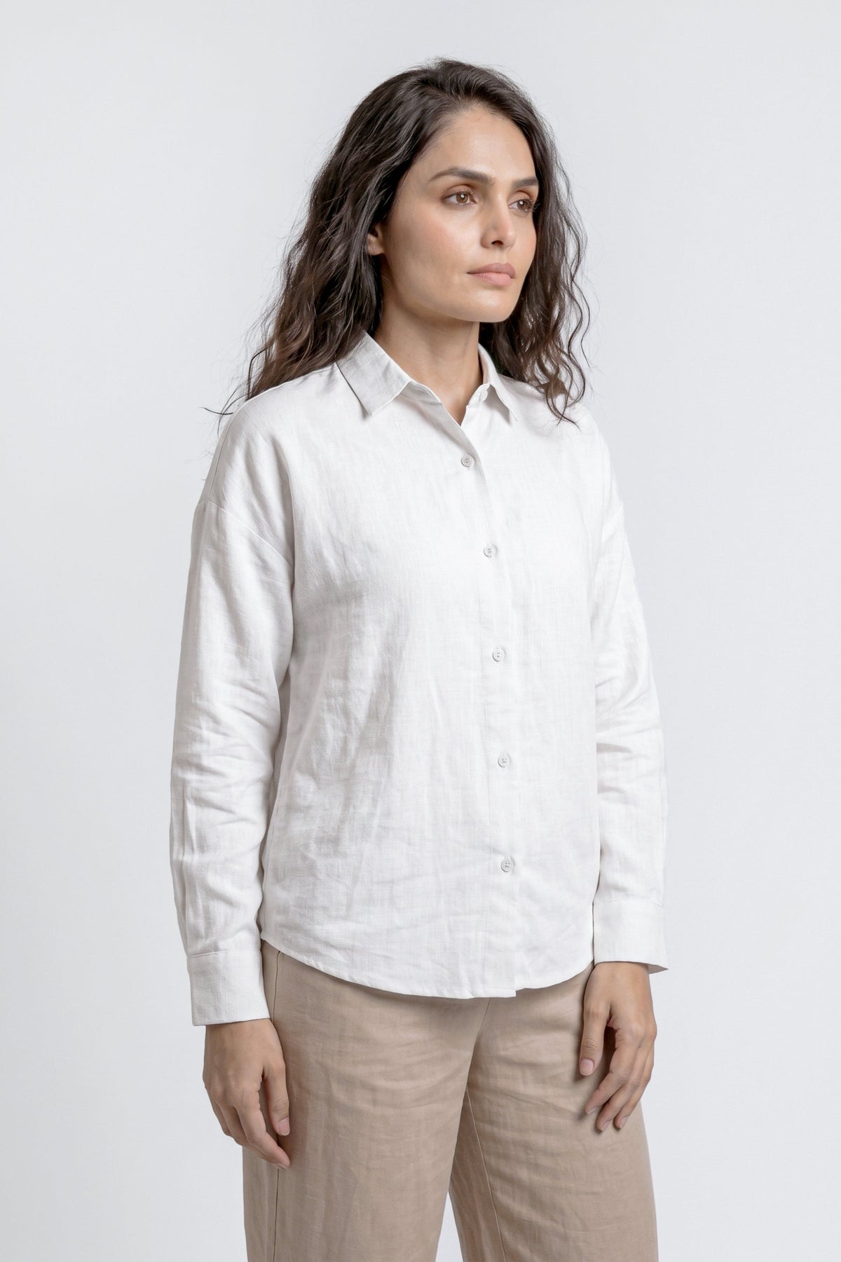 A woman wearing a white long-sleeve linen button-up shirt and beige trousers, standing against a light gray studio background.