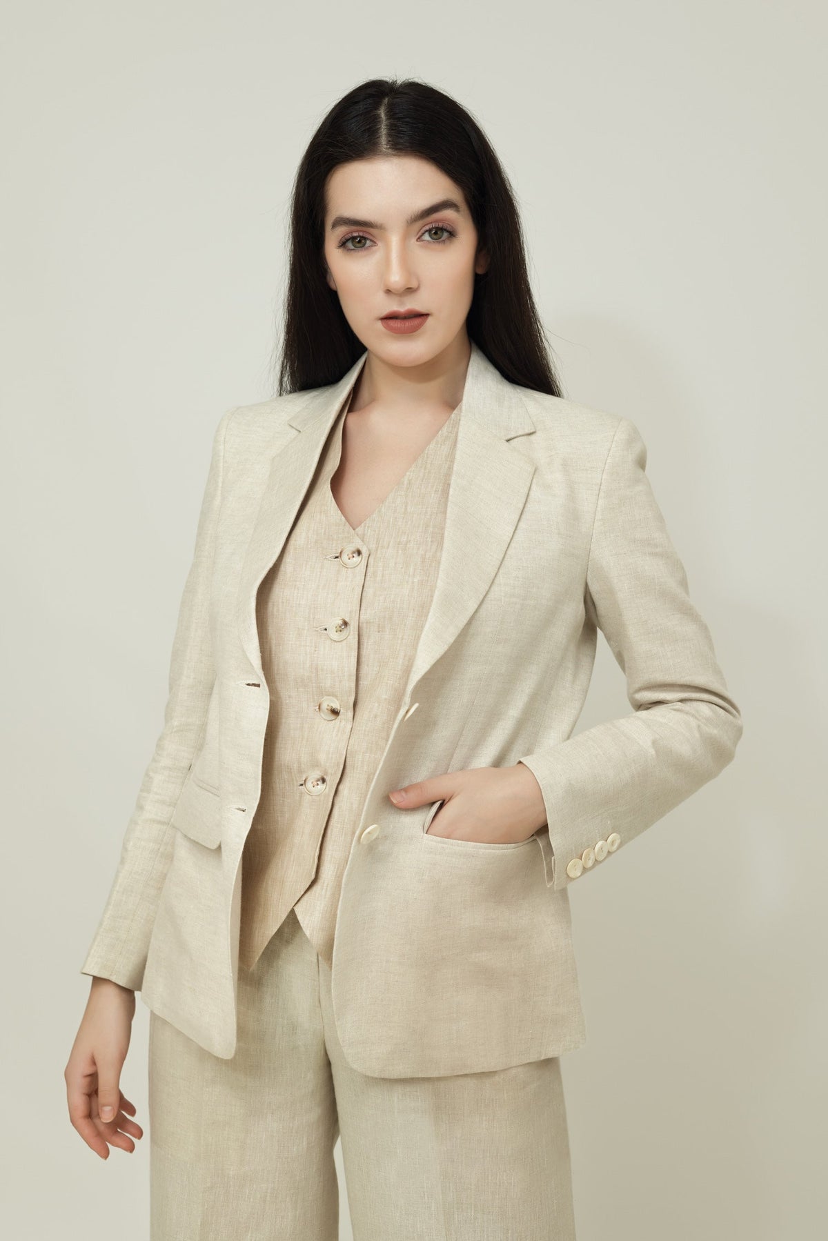 A woman posing against a light gray background, wearing a tailored beige linen three-piece suit. She wears an open single-breasted blazer with notch lapels, a buttoned waistcoat underneath, and matching trousers, keeping one hand resting in her pocket.