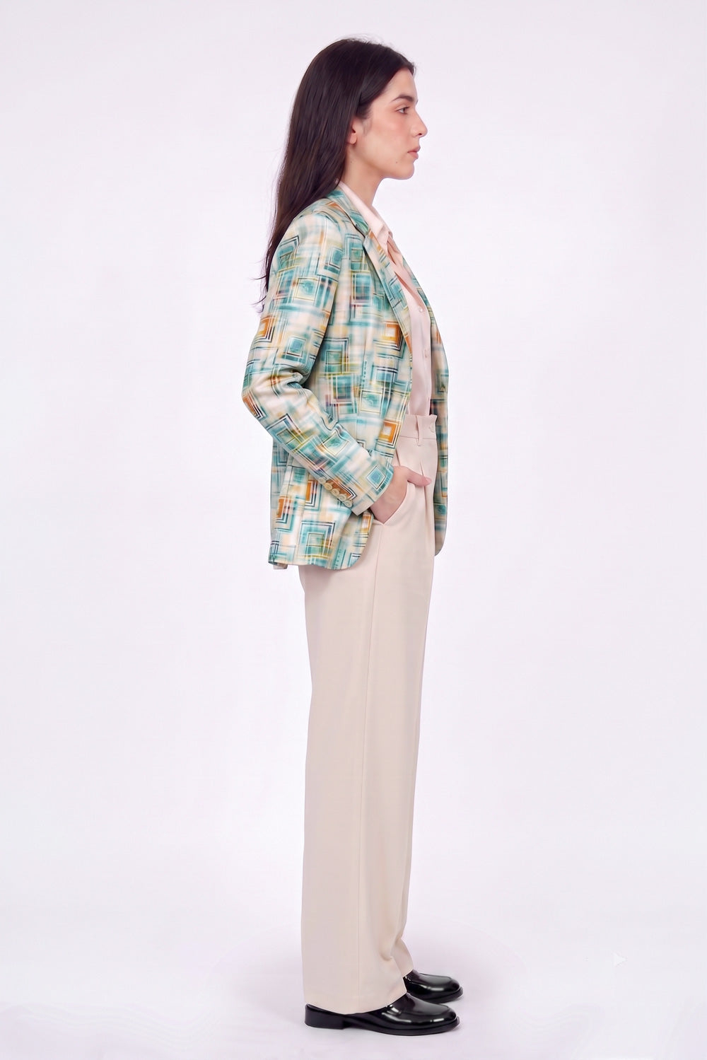 A woman stands in profile wearing a multicolor plaid linen blazer with green, yellow, and blue tones, paired with beige wide-leg trousers and black loafers, against a plain white studio background.
