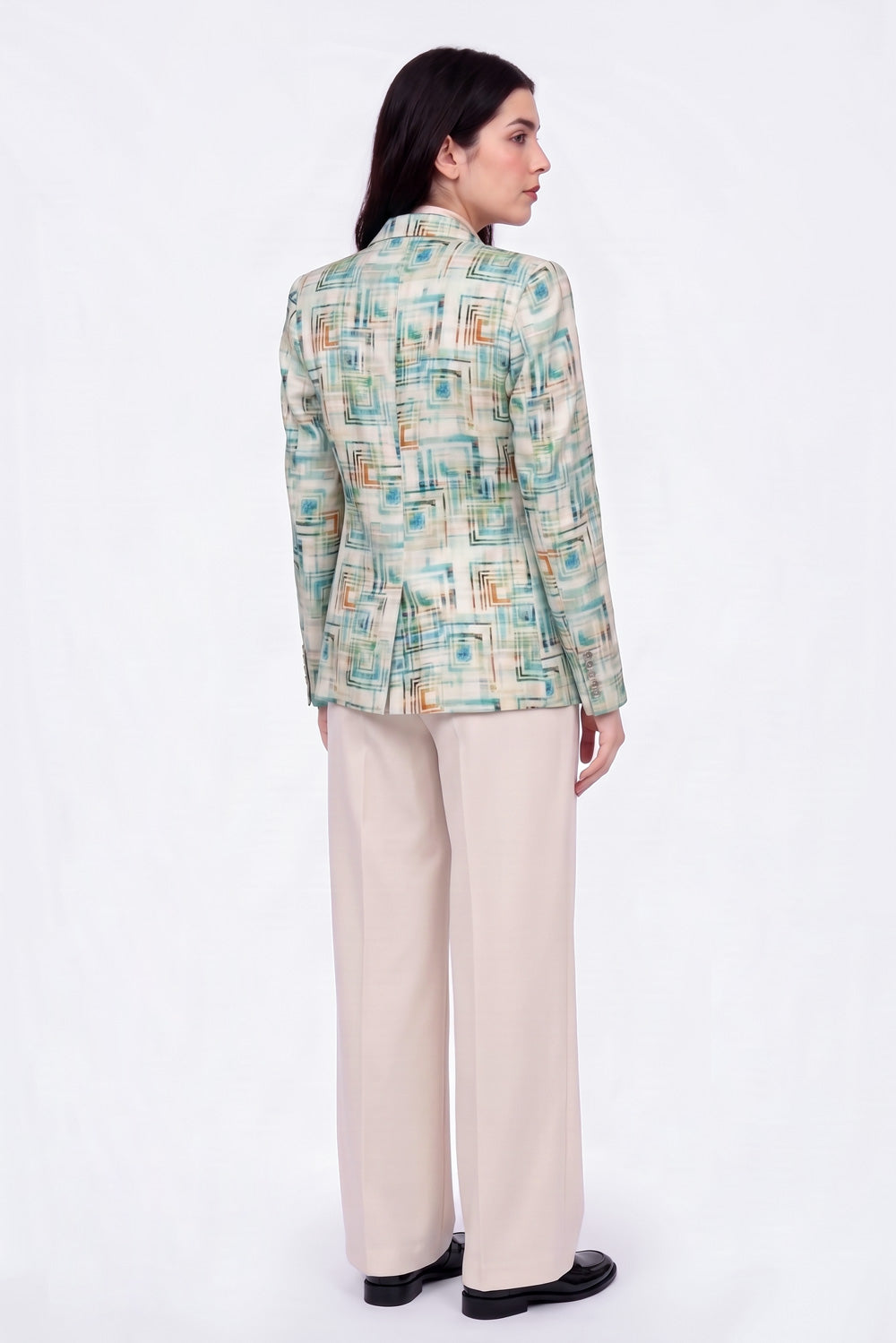 A woman is shown from the back wearing a long-sleeve linen blazer with an abstract geometric pattern in teal, beige, and orange, paired with beige wide-leg trousers and black shoes, against a plain white studio background.