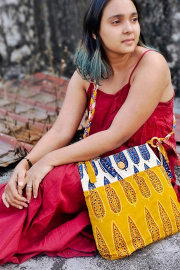 Close-up of a seated woman wearing a sleeveless maroon dress, showcasing a sustainable yellow and off-white ethnic sling bag with intricate leaf block prints and eco-friendly braided detailing.