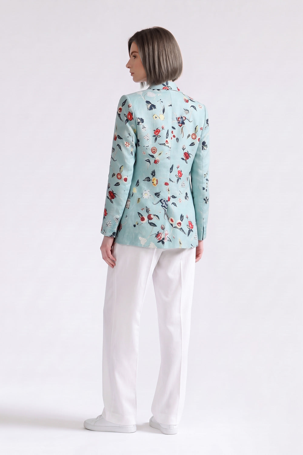 A woman shown from the back wearing a light blue floral linen blazer and white trousers, standing against a plain white studio background.