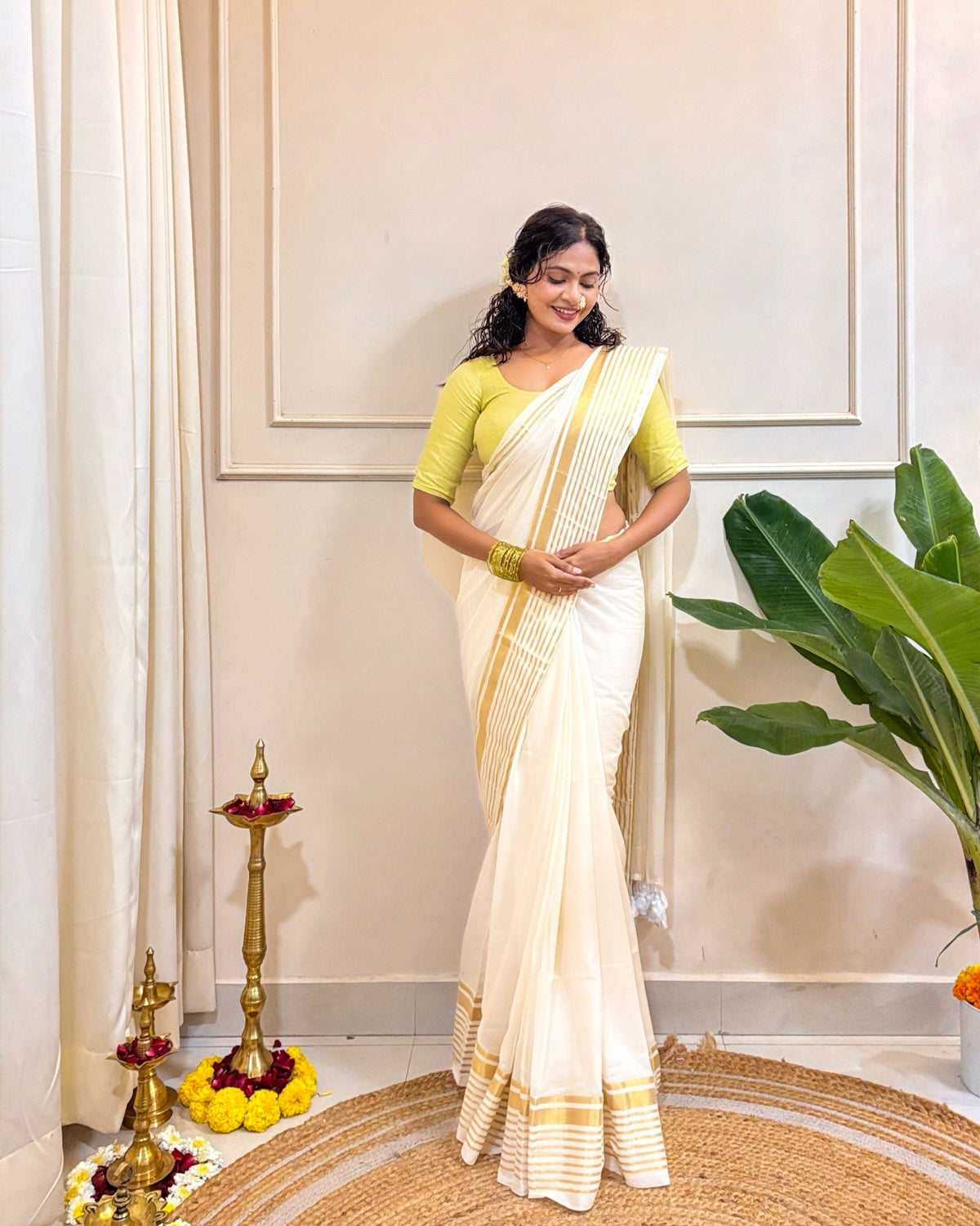 Woman in a white mulmul cotton saree with gold border standing indoors with decorative elements.