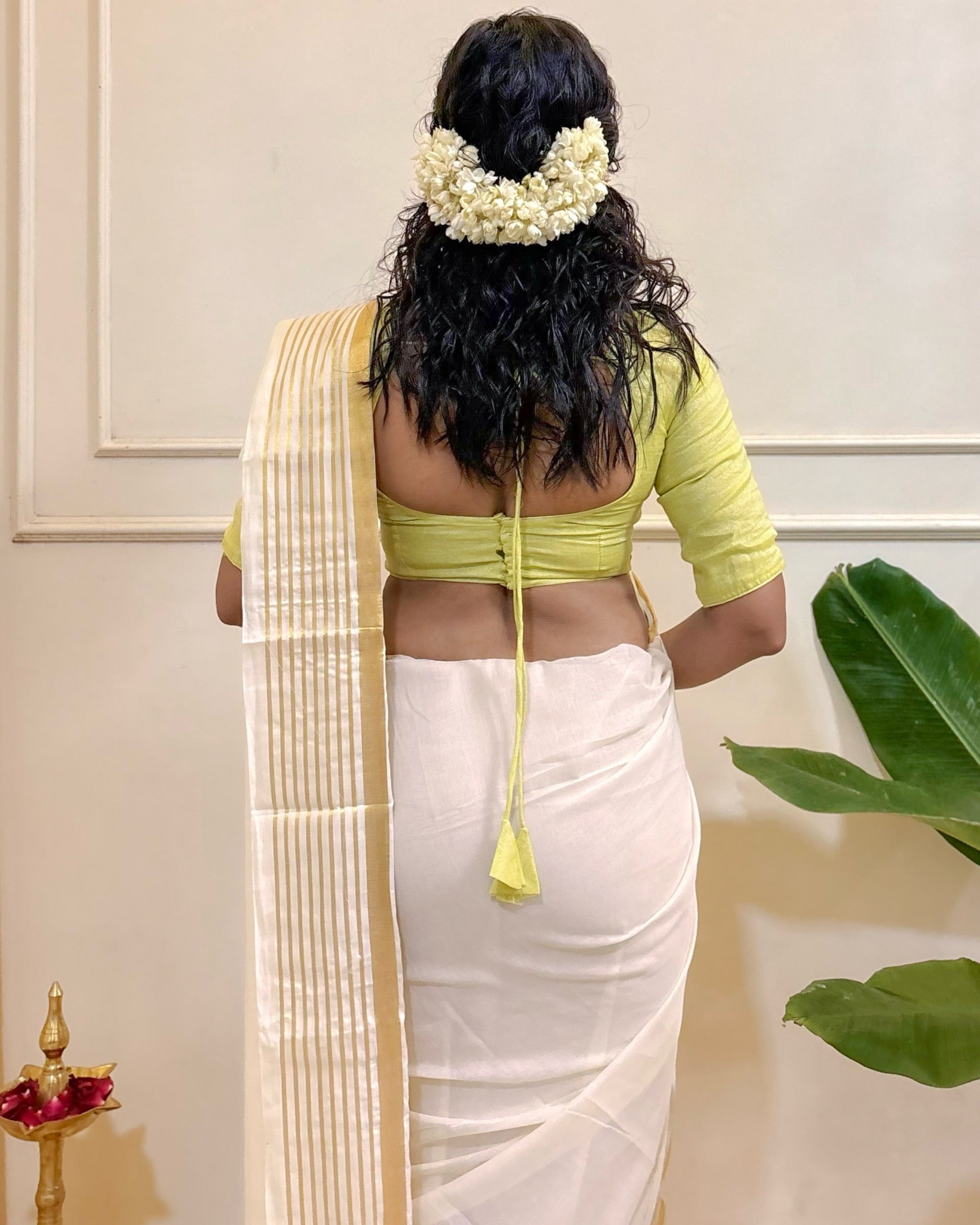Woman in a green blouse and white mulmul cotton saree with floral hair accessories, standing against a neutral background.