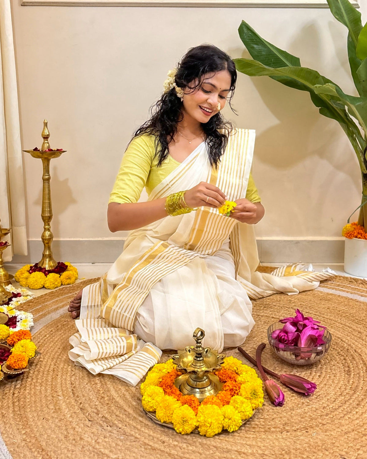 Woman in white mulmul cotton saree arranging flowers on a mat with decorative items around her.
