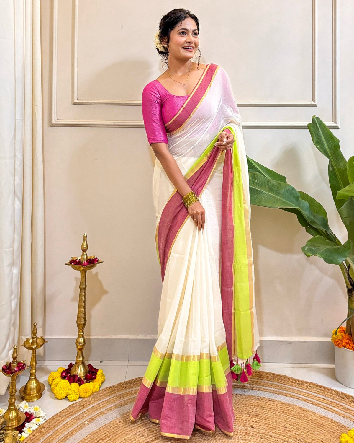Woman in a white mulmul cotton saree with pink and green border standing indoors.