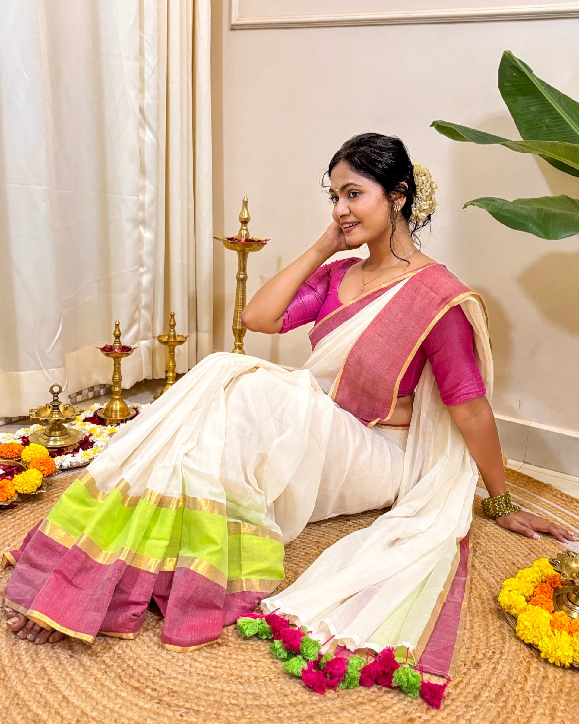 Woman in a white mulmul cotton saree with pink blouse sitting on a mat, surrounded by decorative items.