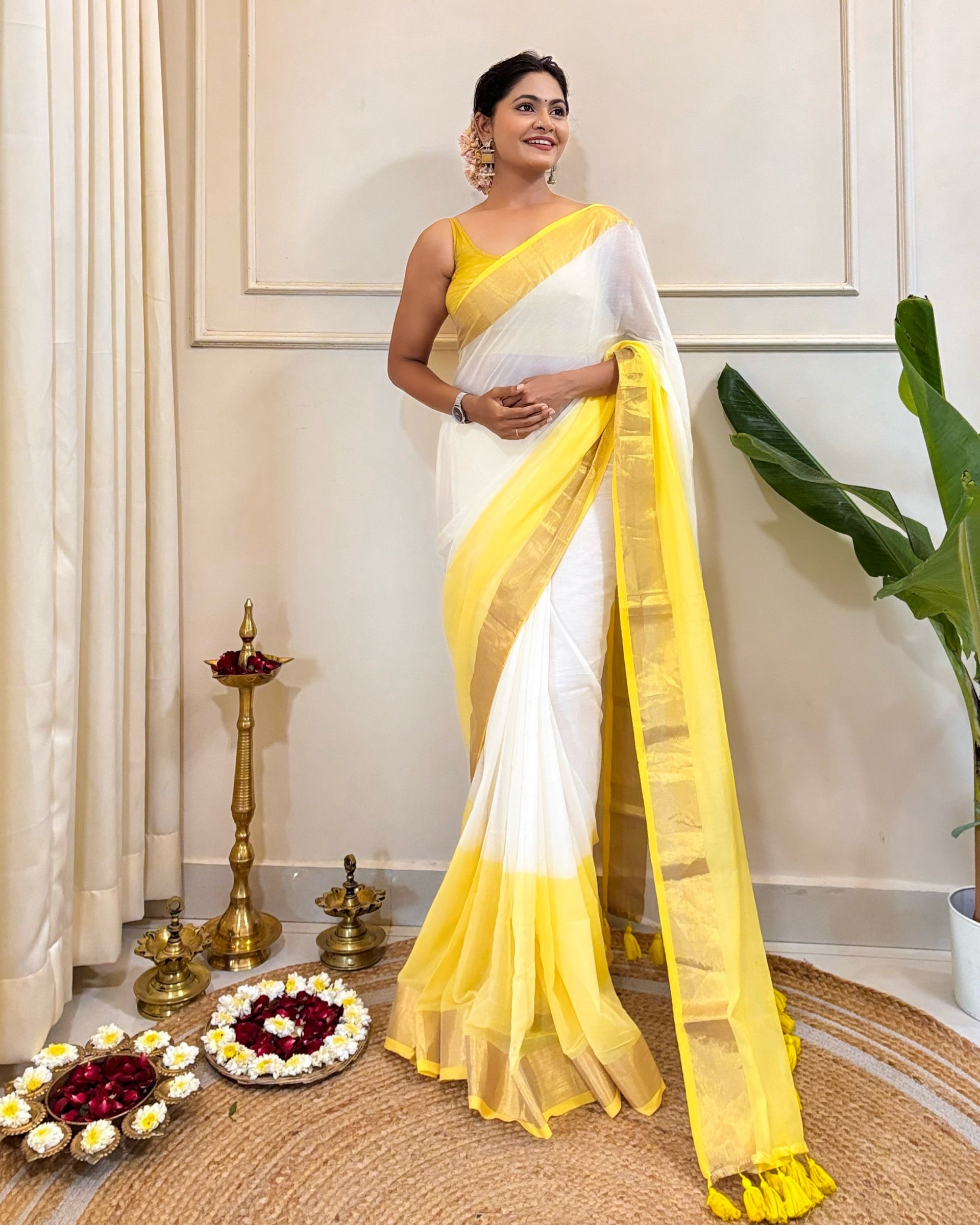 Woman wearing a white and yellow cotton saree in an indoor setting with decorative items.