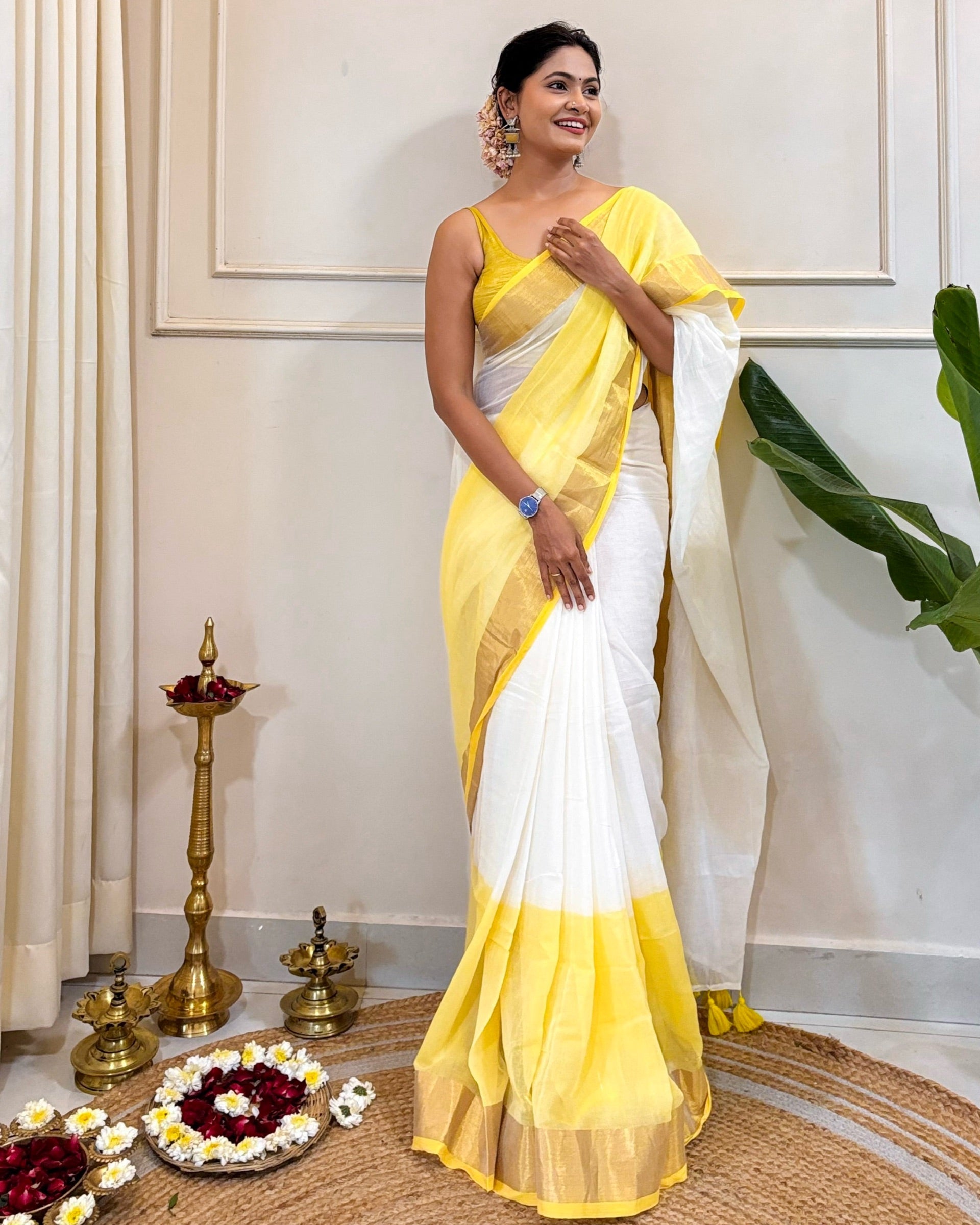 Woman in a yellow and white saree standing indoors with decorative items on the floor.