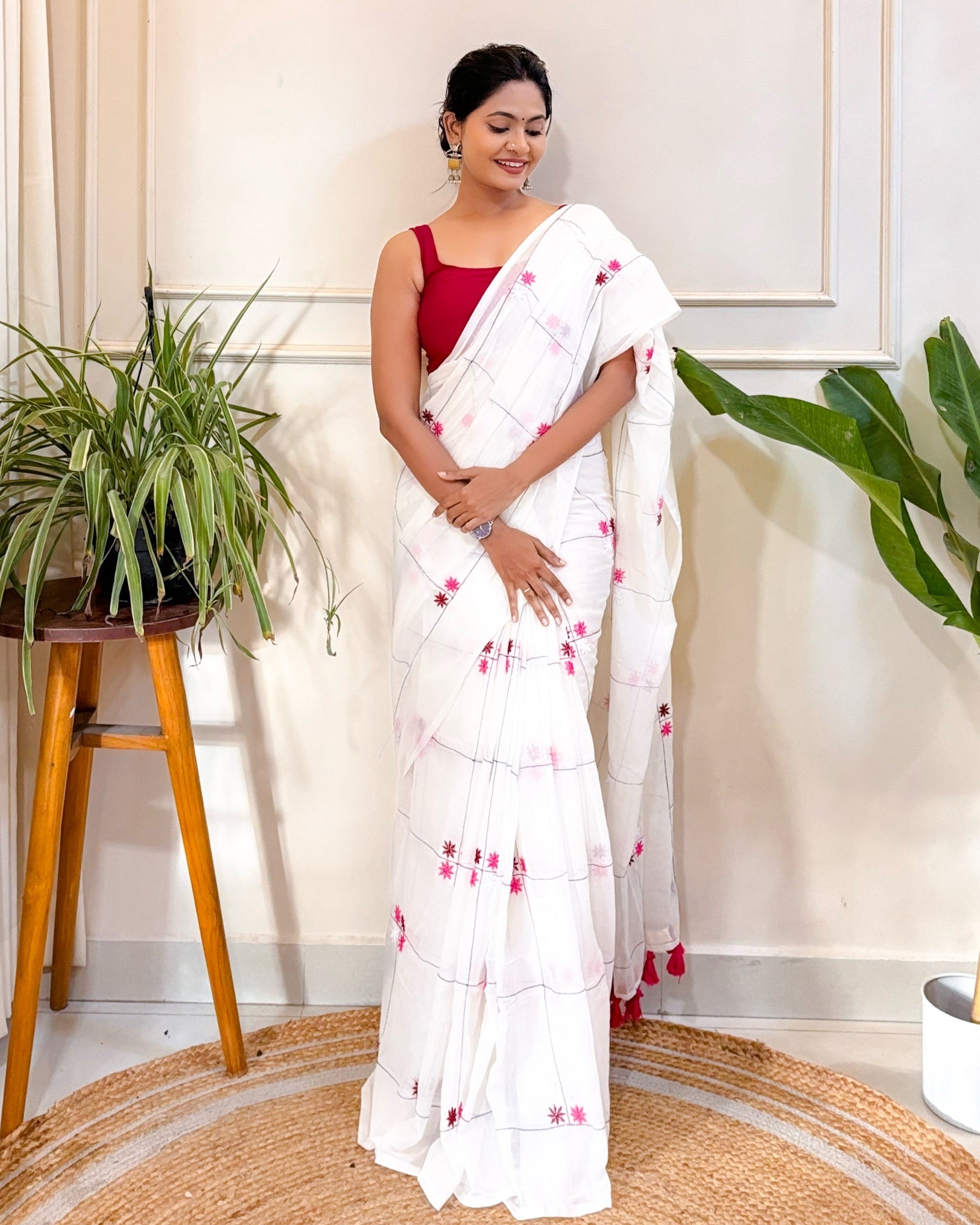 Woman in a white mulmul cotton saree with red patterns standing indoors with plants around.