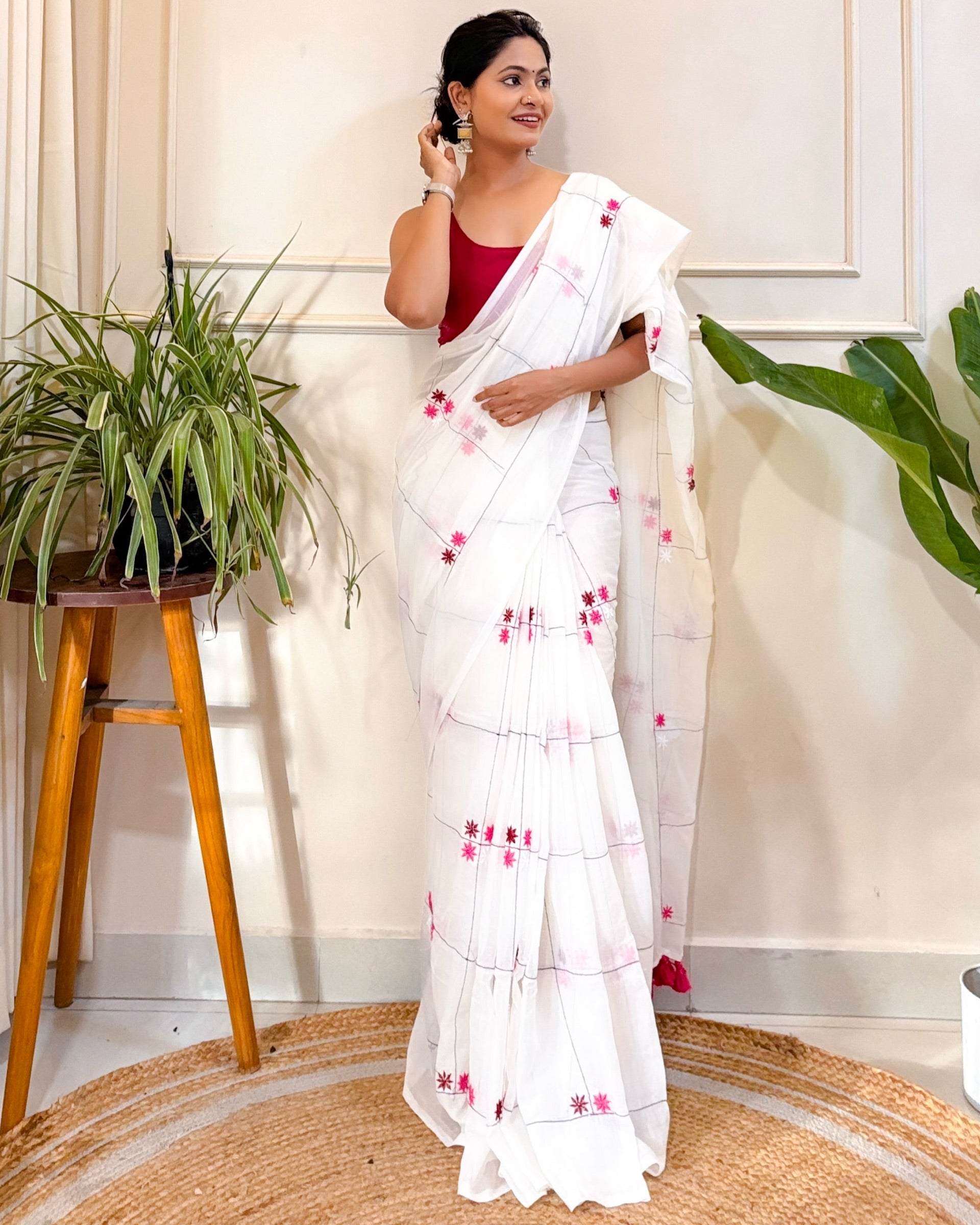 Woman wearing a white mulmul cotton saree with red patterns in a room with plants and a stool.