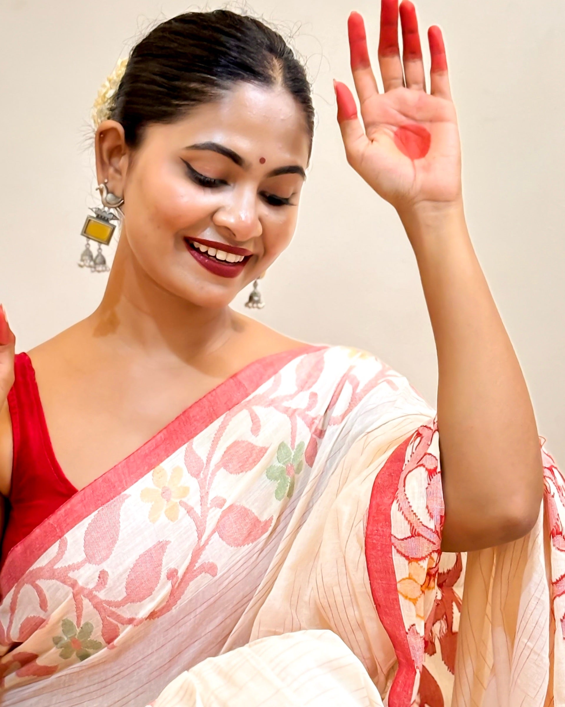 Woman in a traditional mulmul cotton saree with red and beige colors, smiling and showing her hand.