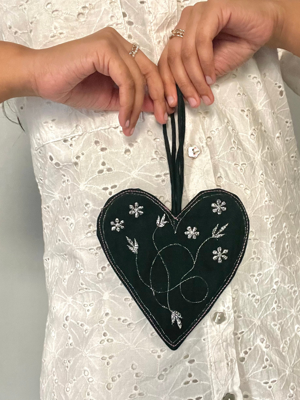 A heart-shaped black fabric bag with metallic thread embroidery, held in hands against a white lace background.