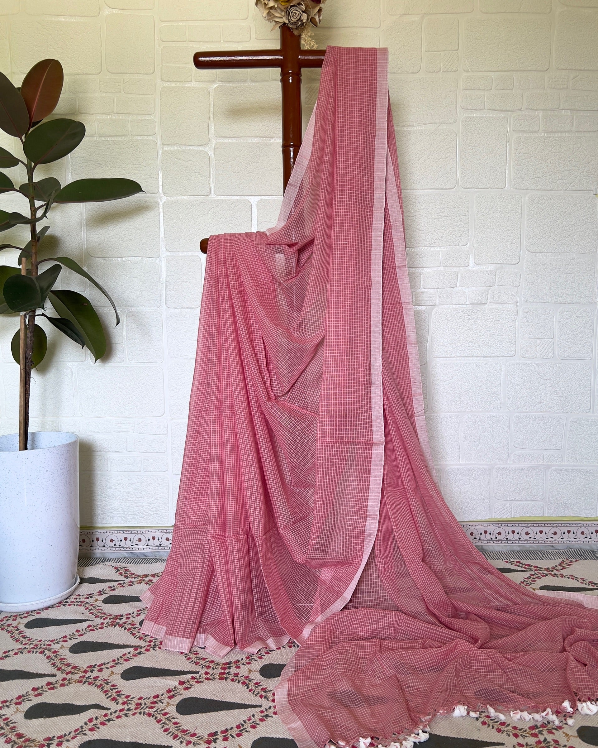 Pink handloom cotton saree draped over a wooden rod against a white brick wall.