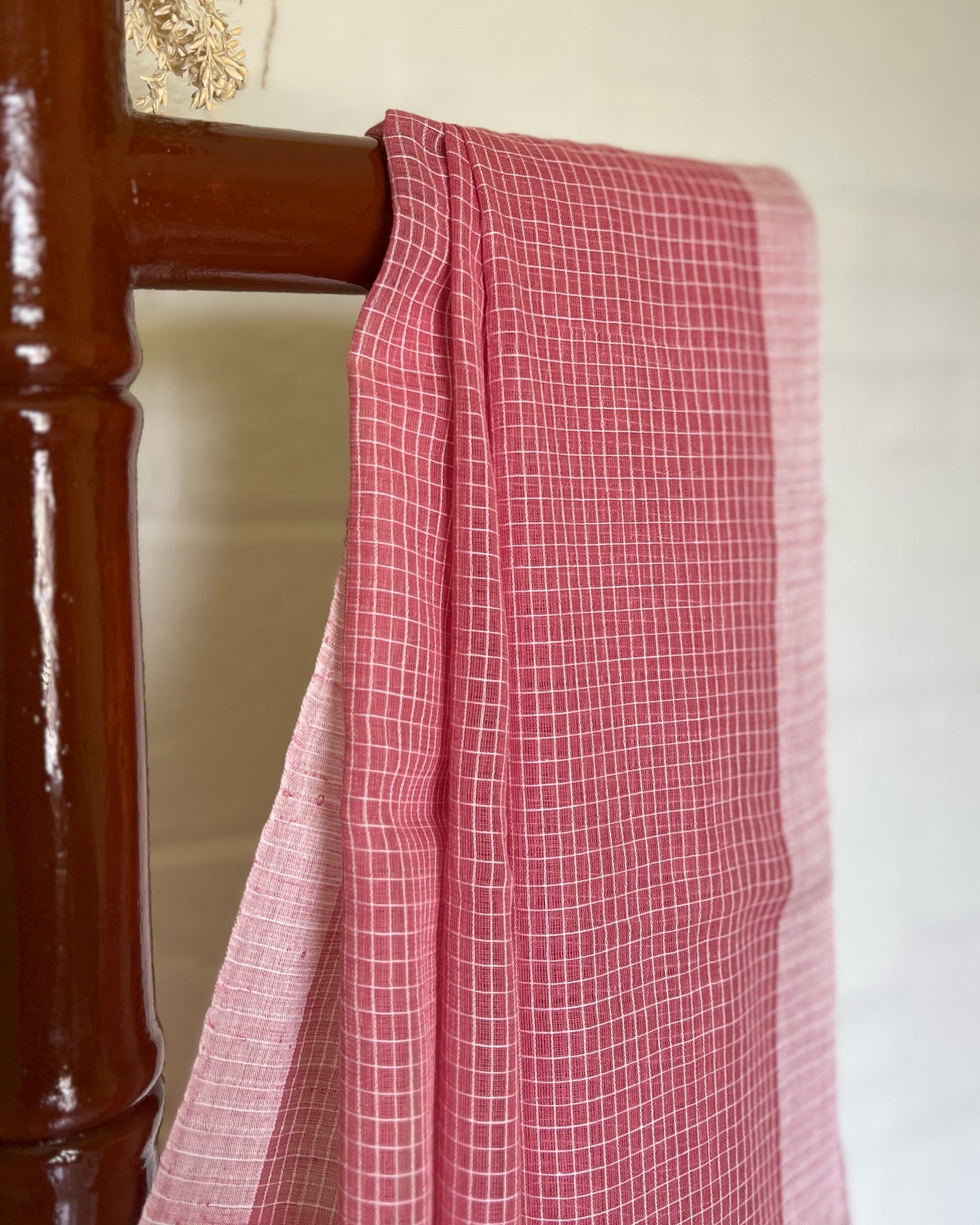 Pink checkered handloom cotton Shopaame saree draped over a wooden chair against a white wall.