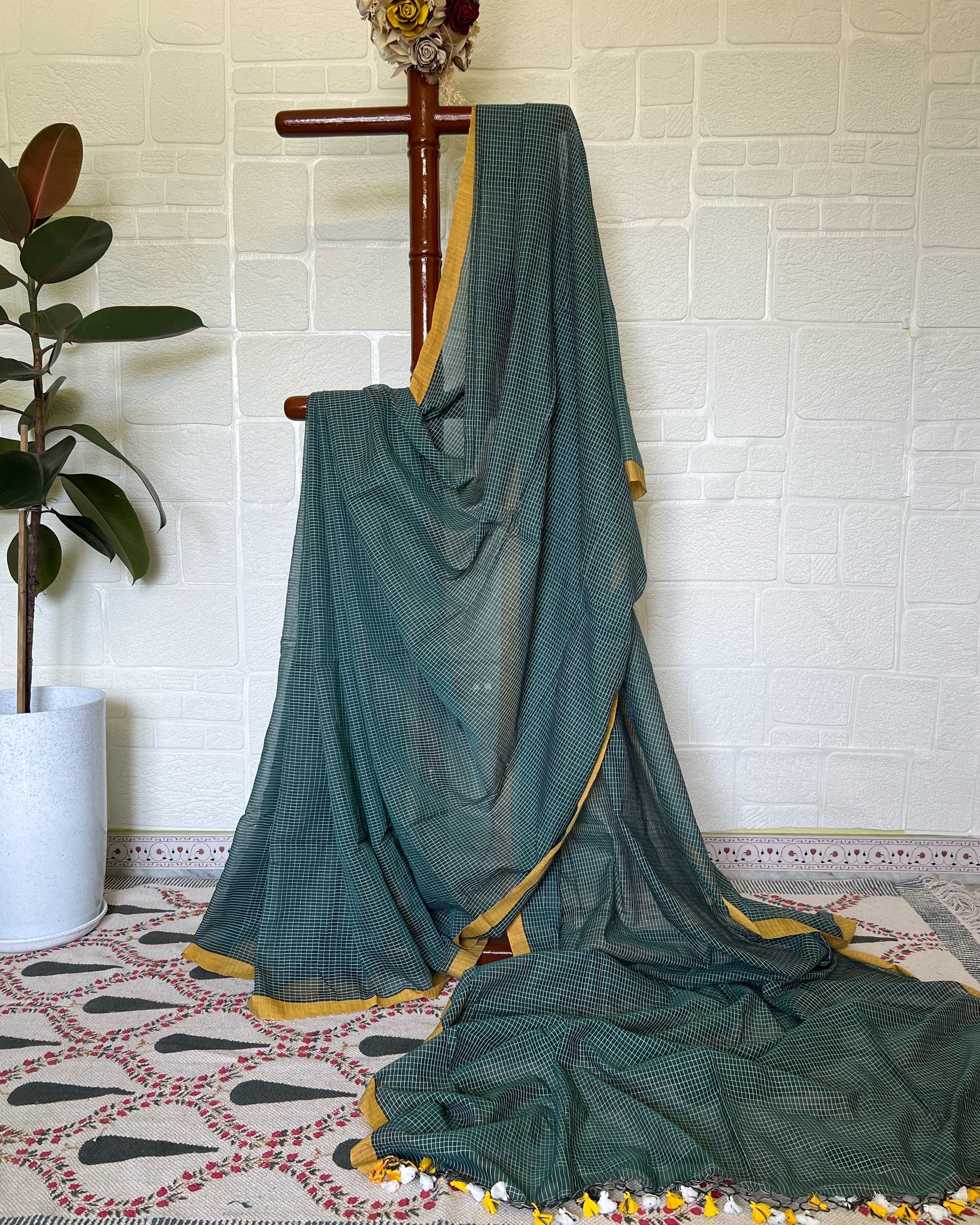 Green check handloom cotton saree draped over a wooden hanger against a white brick wall.