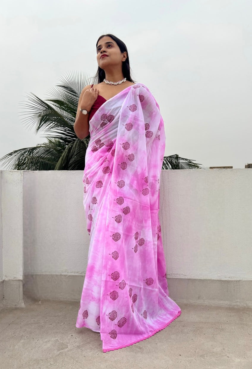 A woman standing on a balcony wearing a pink mulmul cotton saree with floral handblock prints and lace detailing at the aanchal end.