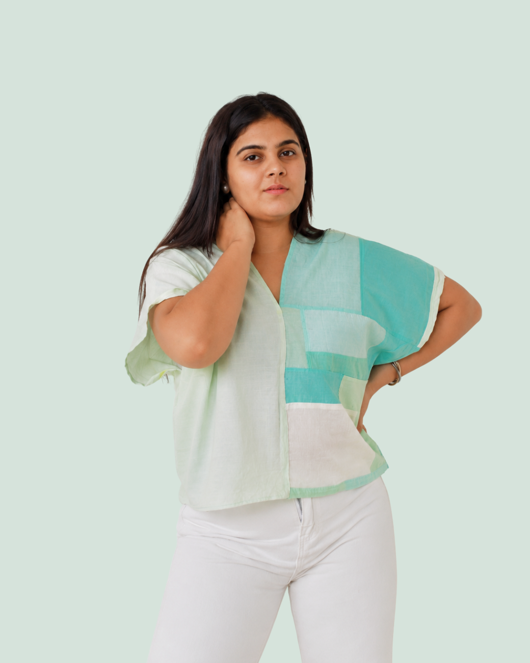 A woman modeling a short-sleeve V-neck kaftan with a patchwork design in mint green, white, and light blue, paired with white pants, against a solid pale green background.
