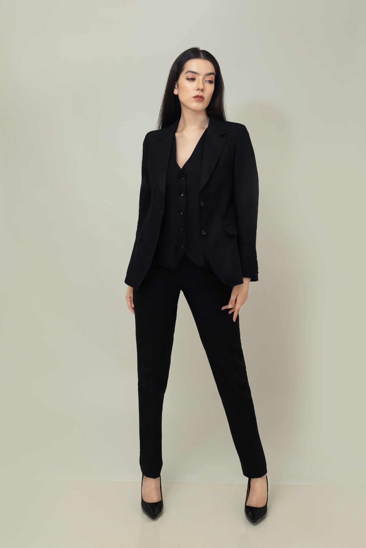 A woman posing against a light gray background, wearing a tailored blue linen blazer. She wears an open single-breasted blazer with notch lapels, a white shirt underneth, and white trousers, holding the blazer with both of her hands.