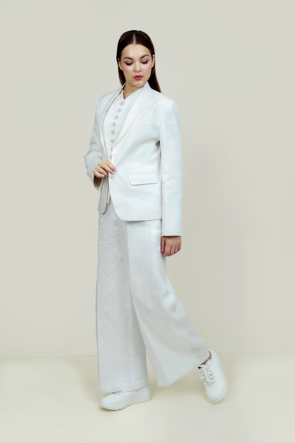 A woman posing against a light gray background, wearing a tailored white linen three-piece suit. She wears an open single-breasted blazer with notch lapels, a buttoned waistcoat underneath, and matching trousers, keeping one hand resting in her blazer.