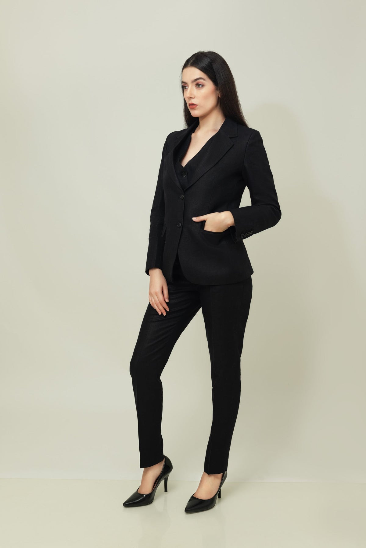 A woman models a tailored black linen blazer with a matching buttoned waistcoat and slim trousers, styled with black pointed heels against a soft neutral studio backdrop. The look is sleek, structured, and contemporary.