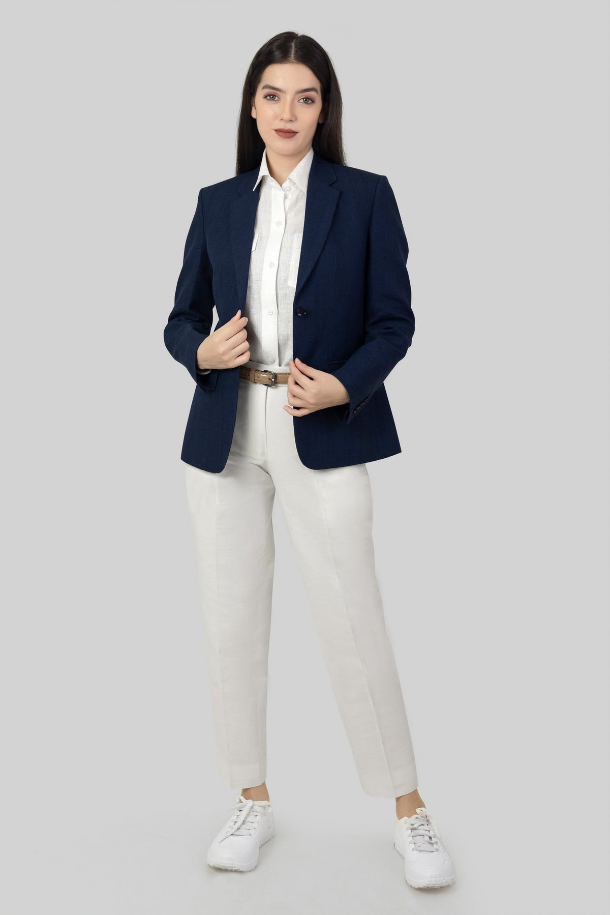A woman posing against a light gray background, wearing a tailored blue linen blazer. She wears an open single-breasted blazer with notch lapels, a white shirt underneth, and white trousers, holding the blazer with both of her hands.