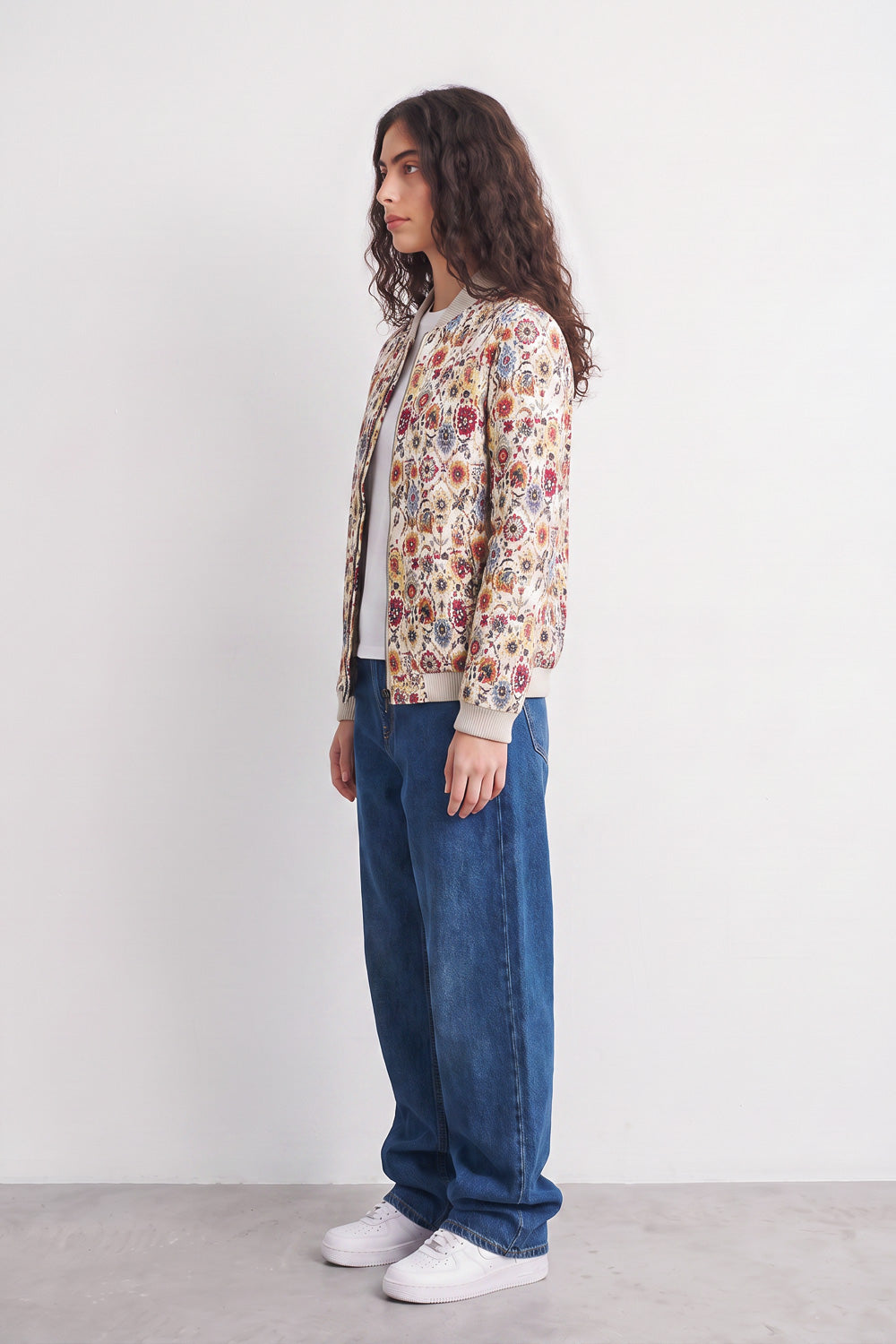 A woman stands in profile wearing a cream-colored bomber linen jacket covered in a dense multicolor floral and botanical print, paired with wide-leg blue jeans and white sneakers, against a plain white studio background.