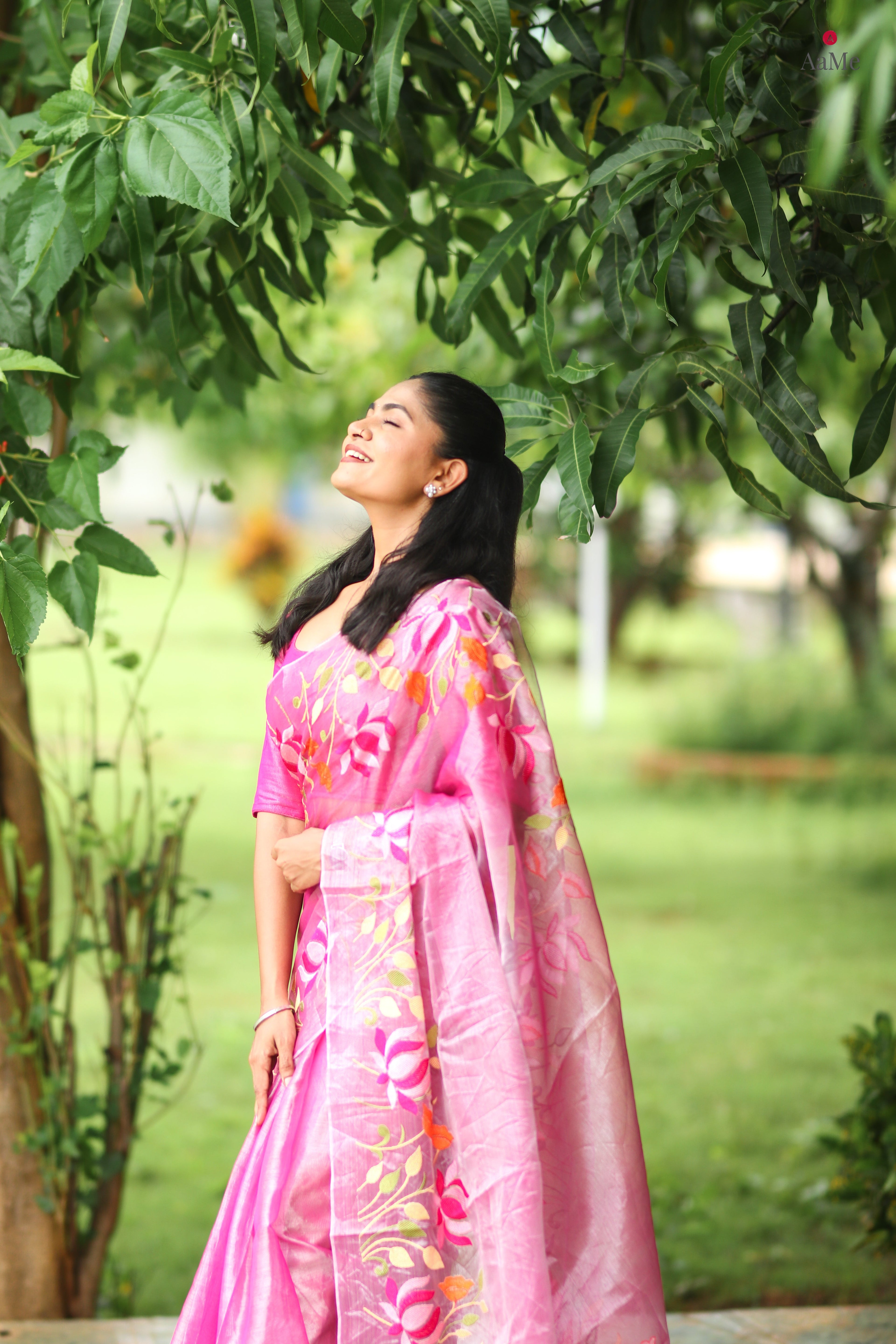 Woman in a pink tissue saree with floral patterns standing under green foliage