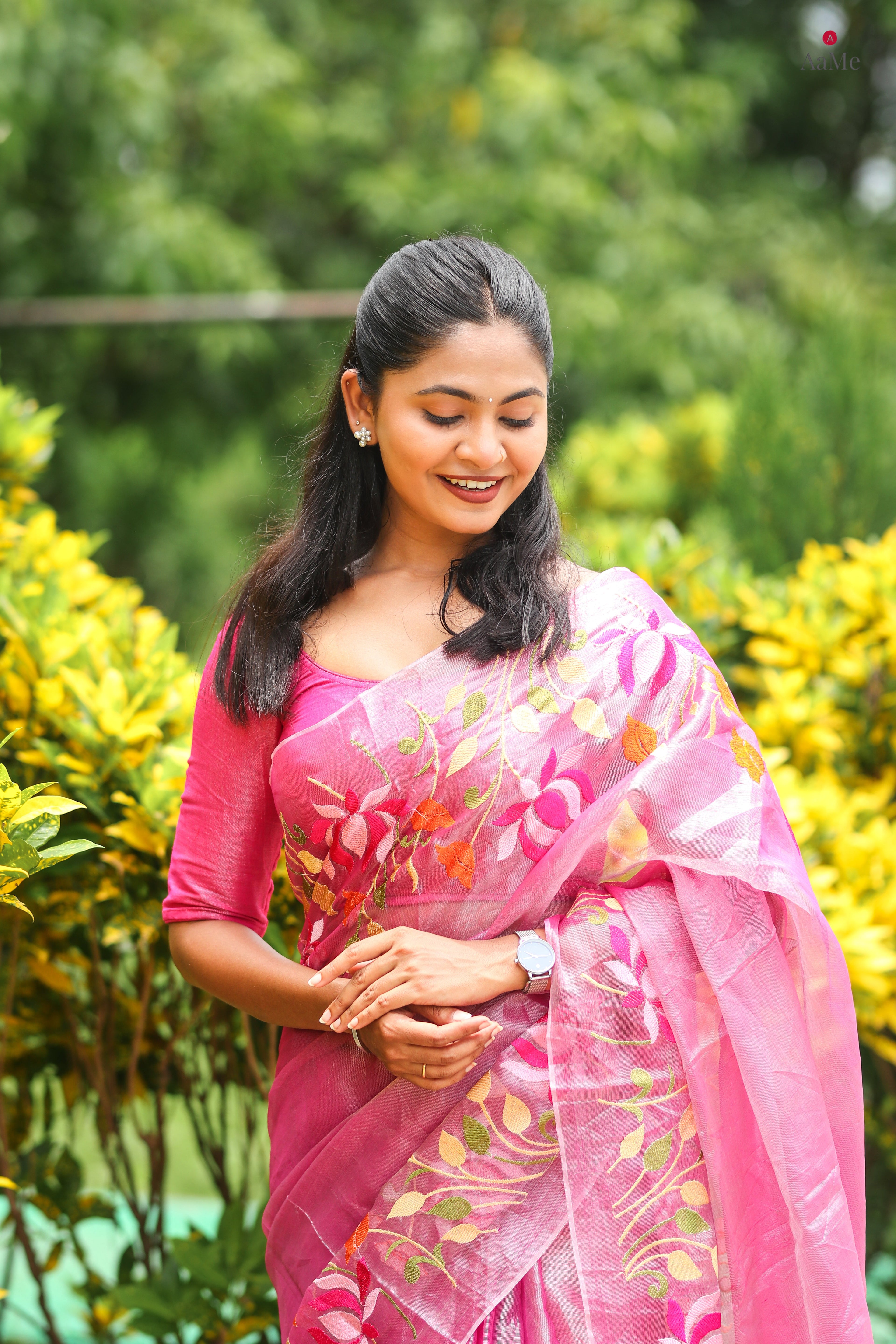 Woman in a pink saree with floral patterns standing in front of green foliage.