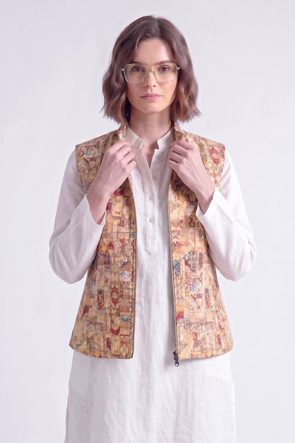 A woman wearing a white shirt and a beige linen vest with a colorful, detailed traditional pattern, standing against a light gray background.