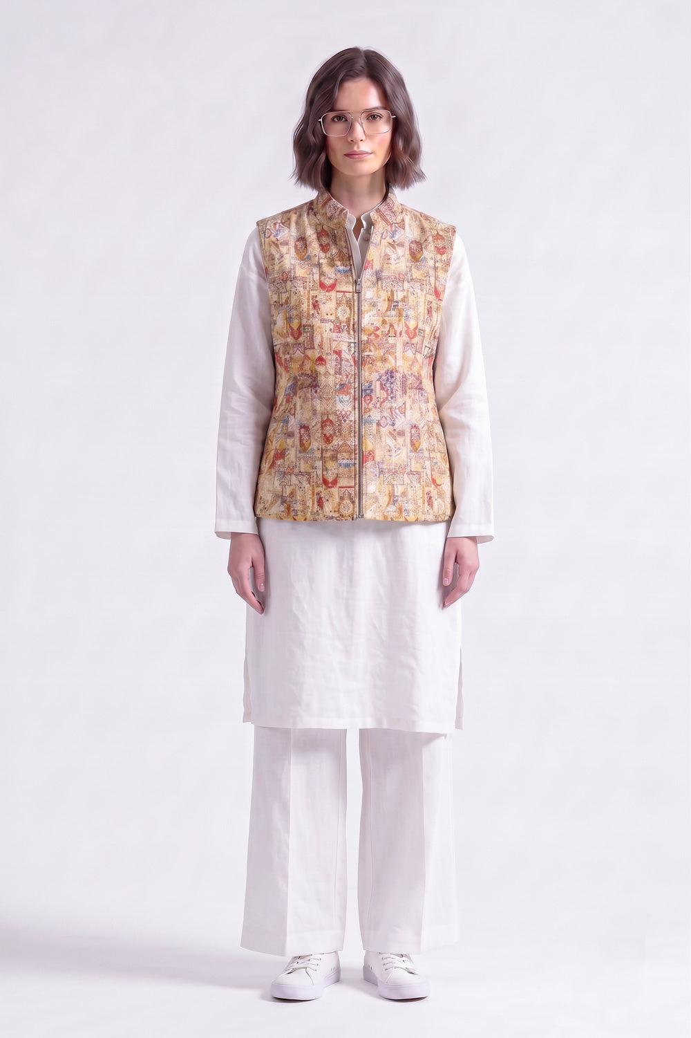 A model wearing a white traditional outfit consisting of a long-sleeve kurta, wide-leg trousers, and white sneakers, layered with a sleeveless linen vest featuring a detailed multicolor illustration pattern.
