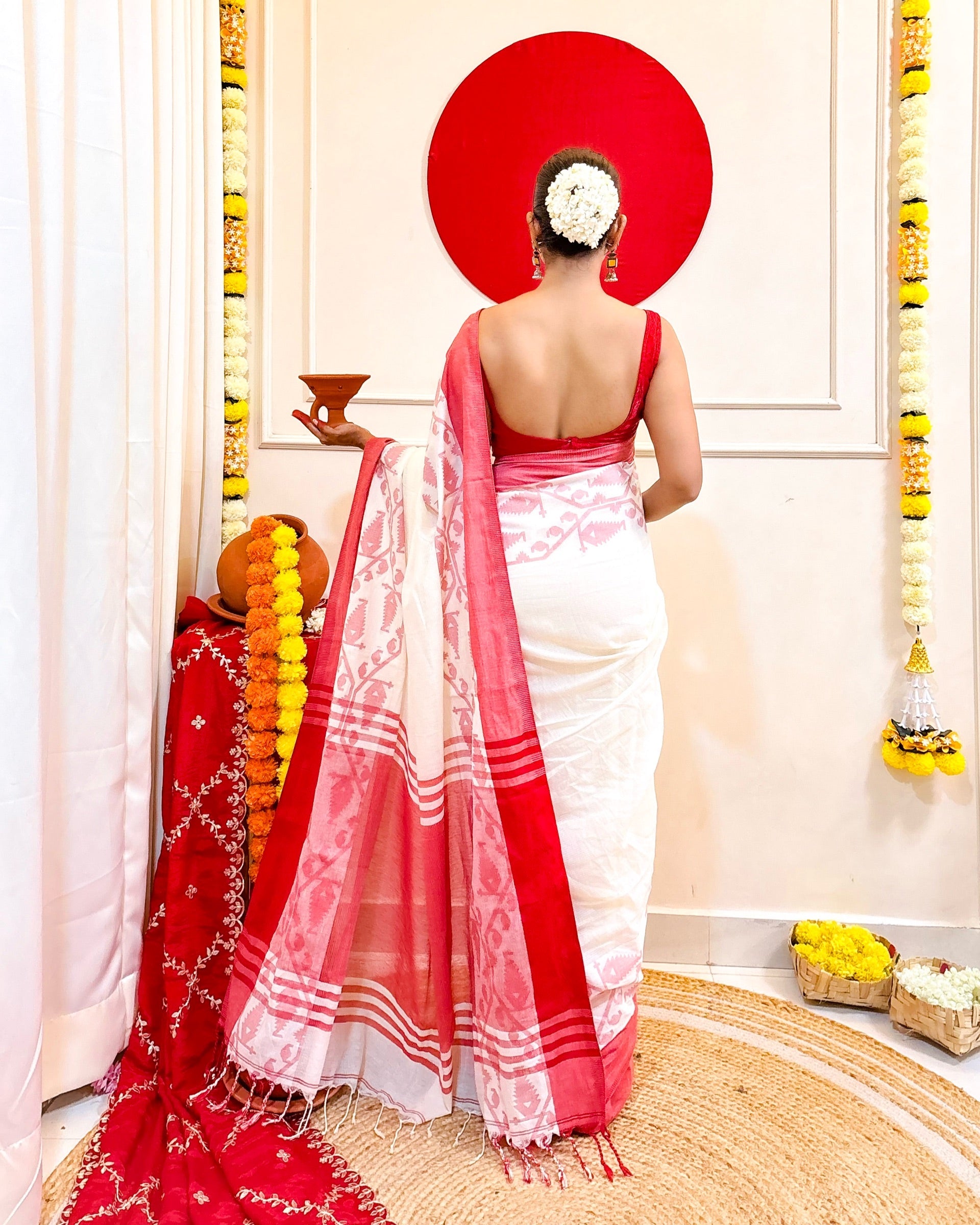 Woman in a white mulmul cotton saree with red border standing in a decorated room.