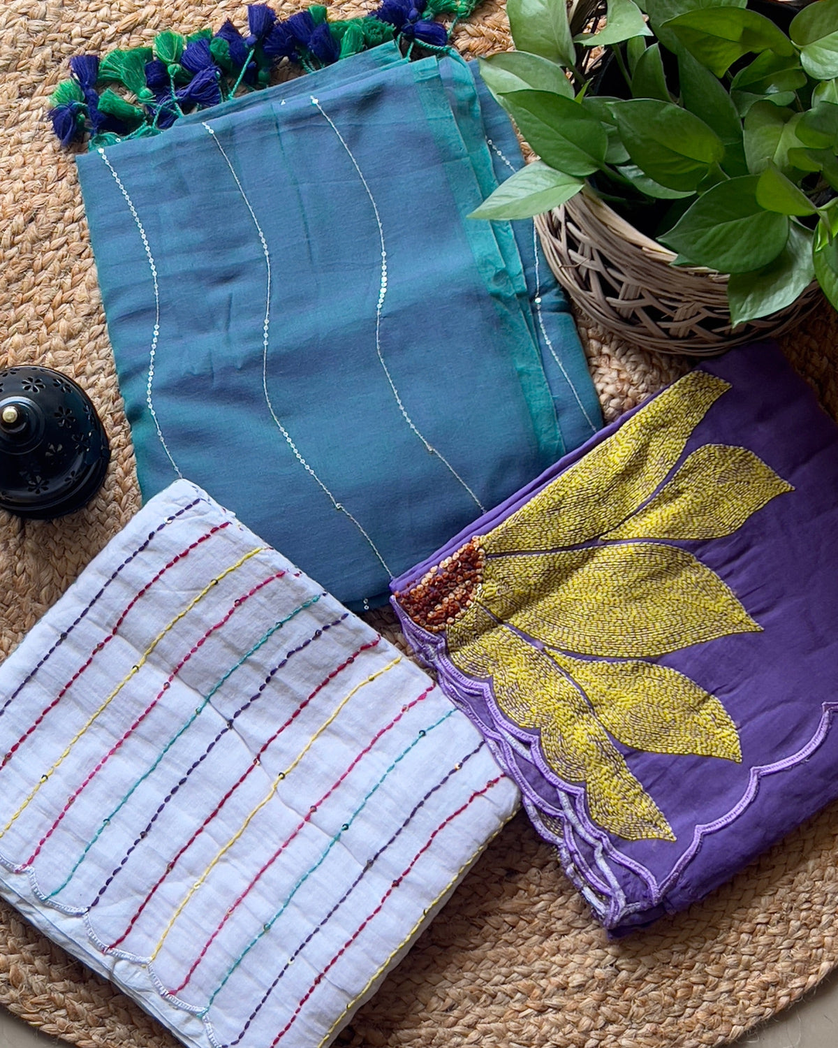 A set of three folded Shopaame sarees displayed on a flat surface with a plant in the background. The sarees are in blue, purple and white colors with patterned designs.