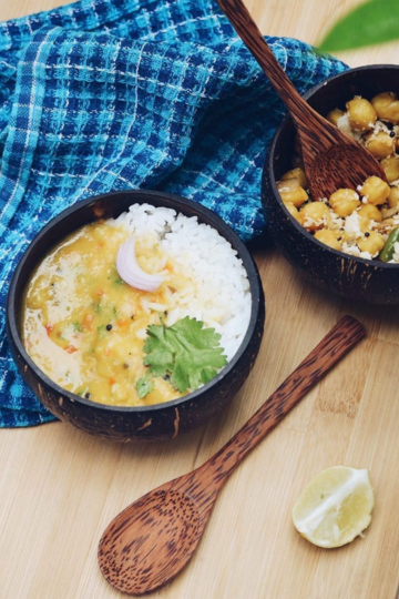 Eco-friendly coconut bowls filled with Indian dal, rice, and chickpeas, served with a wooden spoon on a bamboo surface.