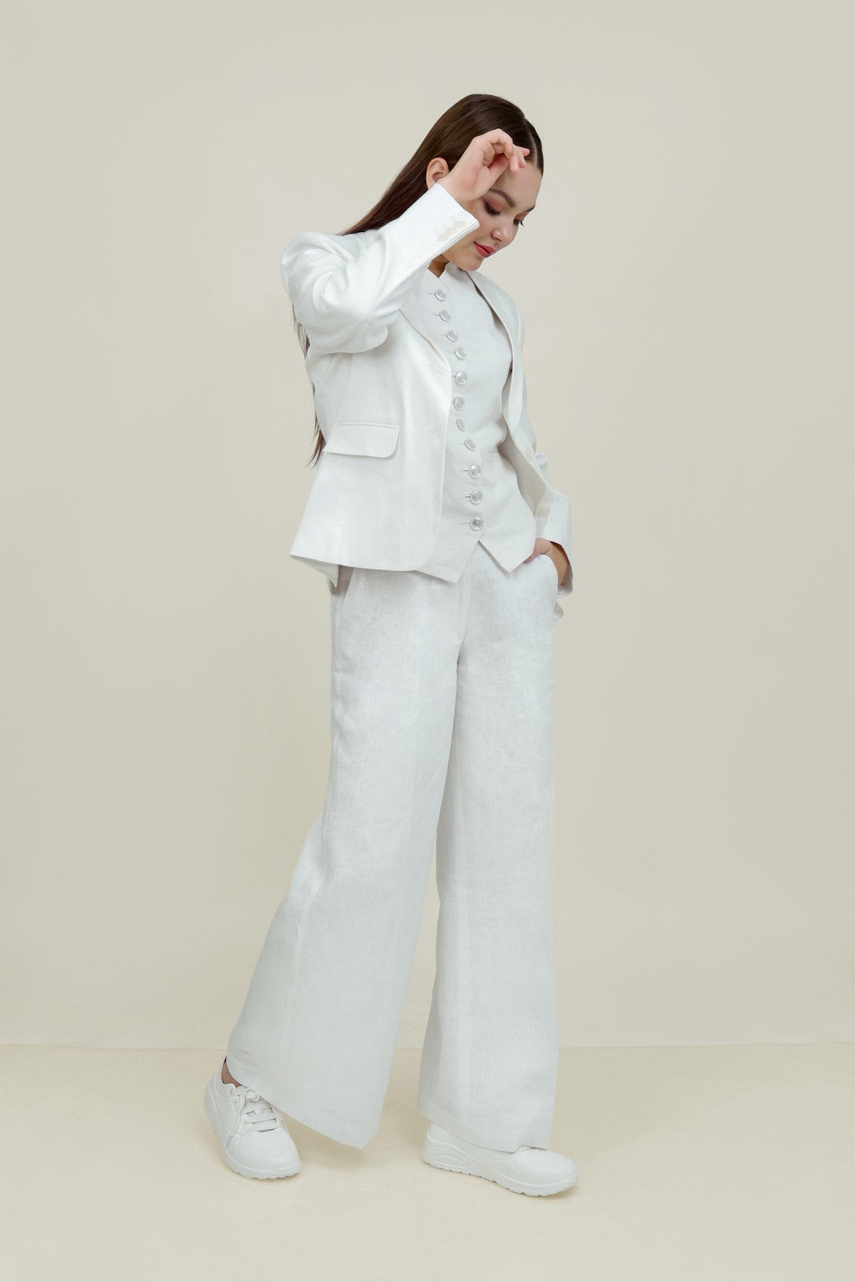 A woman posing against a light gray background, wearing a tailored white linen three-piece suit. She wears an open single-breasted blazer with notch lapels, a buttoned waistcoat underneath, and matching trousers, keeping one hand resting in her pocket.