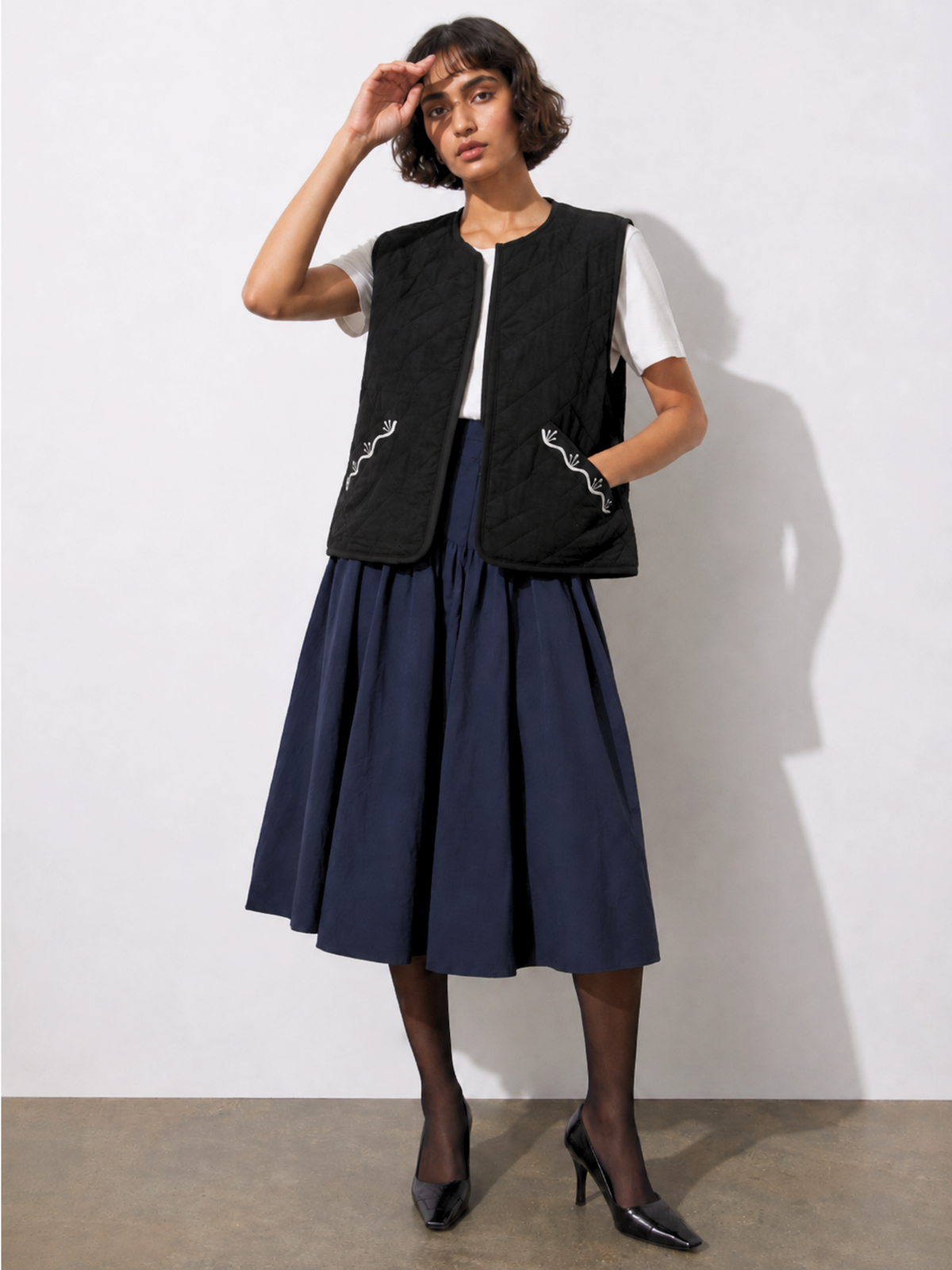 a model wearing black embroidered half quilted jacket with a white shirt and navy skirt against a white background.