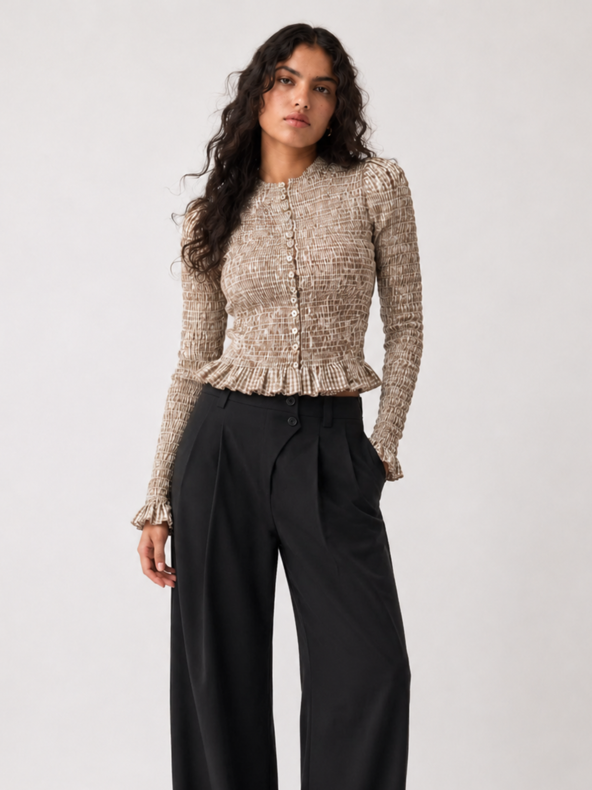 Woman wearing a brown and white top with handcrafted smocking detailing and black pants on a white background