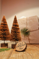 Two medium-sized handcrafted coir Christmas trees from Almitra Sustainables displayed on a rustic wooden table, with a fabric-wrapped gift box and wooden "Oh Christmas Tree" tag in the background.