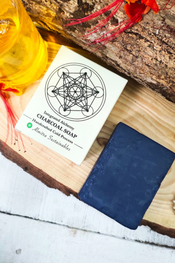 Almitra Sustainable activated charcoal cold process soap bar beside natural oil bottle on wooden surface, handmade detox skincare soap.