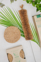Flat lay of a sustainable kitchen cleaning combo featuring a round coconut coir scrub pad and long bottle brush with coir bristles, placed on palm leaves alongside eco-conscious packaging, representing Almitra Sustainables’ natural zero-waste cleaning products.