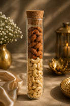 A clear glass test tube with a cork stopper, filled with whole almonds on top and cashews below, styled on a beige fabric surface with warm decorative props in the background.