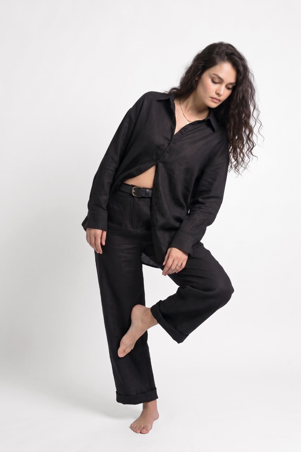 A woman modeling a black long-sleeve button-up linen shirt and matching black wide-leg pants against a plain light gray studio background.