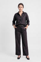 A model stands against a light gray studio background wearing a black long-sleeve button-up linen shirt, black wide-leg trousers, a black belt and black heels.