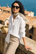 A woman wearing a white linen button-up linen shirt, beige trousers, a beige patterned scarf, and black sunglasses, leaning on a stone wall with a Mediterranean town and blue sea in the background.