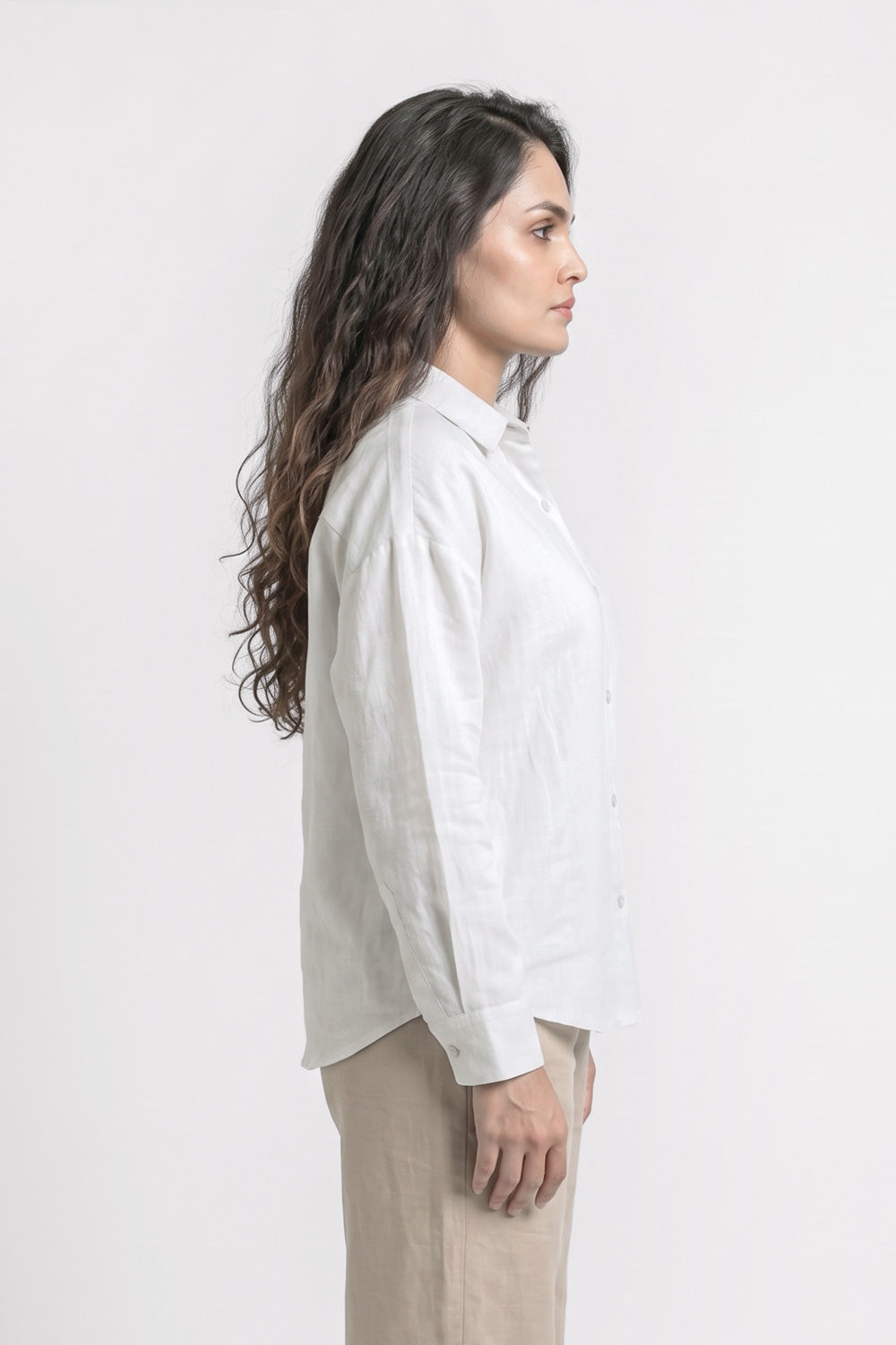 A woman shown in profile wearing a white long-sleeve linen button-up shirt and beige trousers, photographed against a clean white studio background.